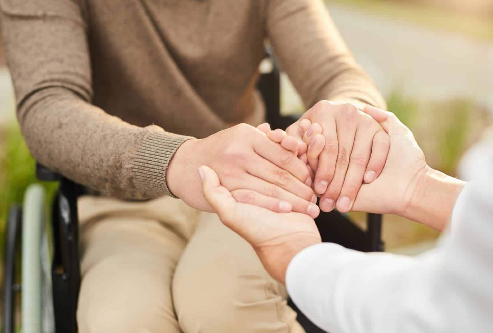 אדם בכיסא גלגלים מחזיק ידיים באופן תומך ומחבק עם אדם אחר לידו, בסביבה טבעית שטופת שמש, סמל לתמיכה רגשית וסיוע חברתי בביטוח לאומי.