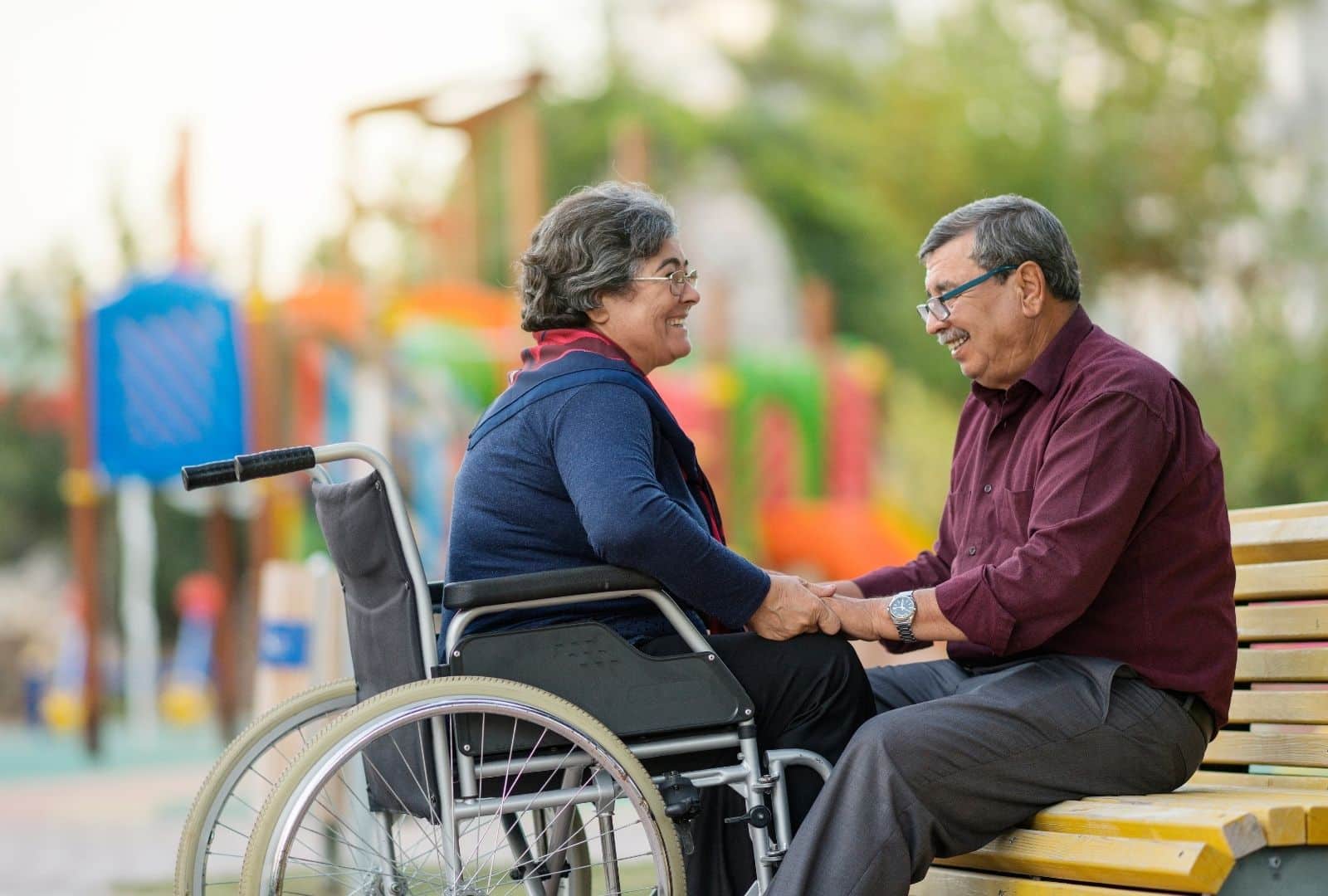 זוג קשישים מחייכים ומחזיקים ידיים בפארק נגיש, אישה בכיסא גלגלים לצד בעלה על ספסל, מסמל תמיכה משפחתית עם סיעוד.