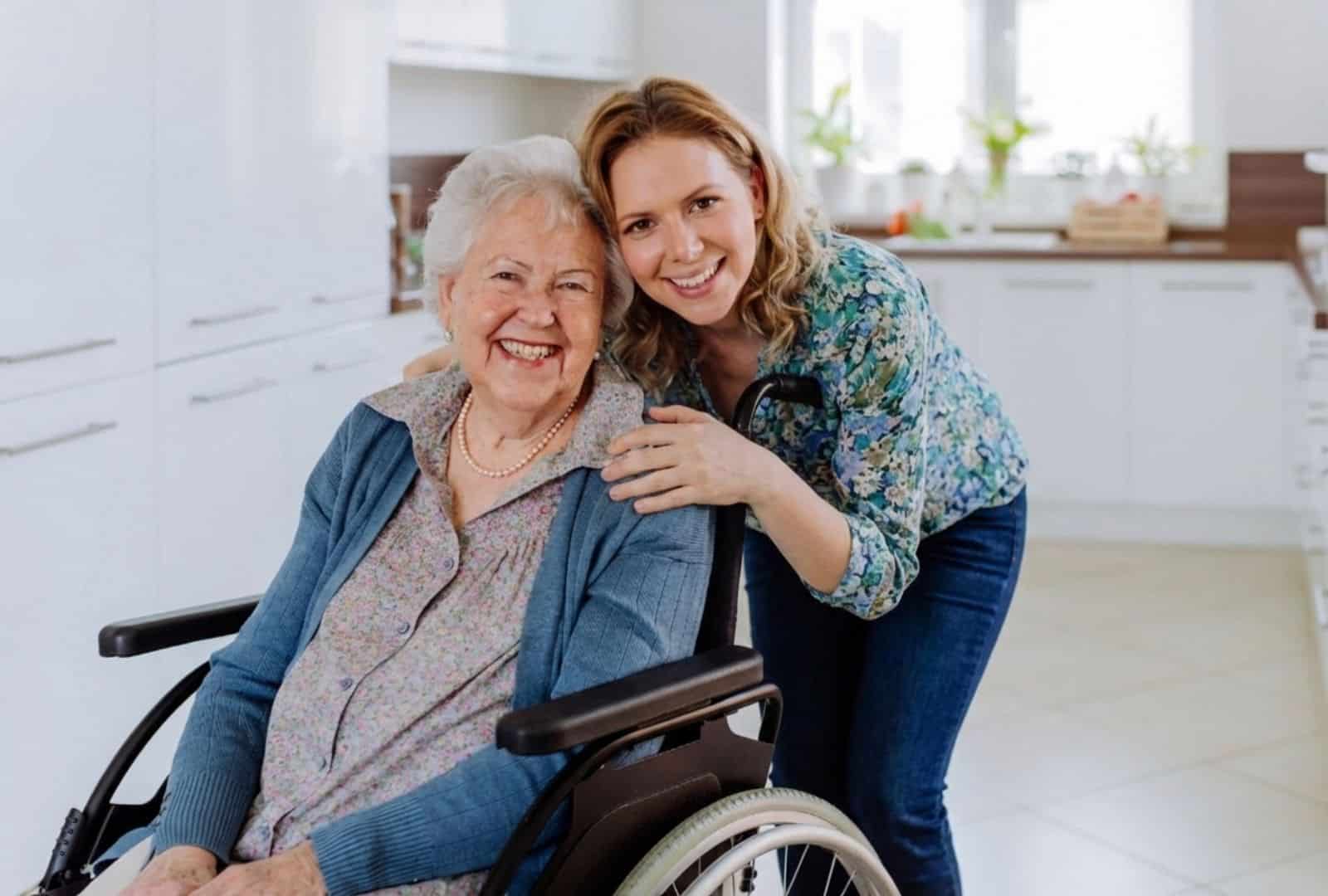 אישה צעירה מחייכת לצד סבתה הקשישה בכיסא גלגלים בבית, ממחישה את הצורך בתו נכה לרכב לקשישים עם מוגבלות ניידות