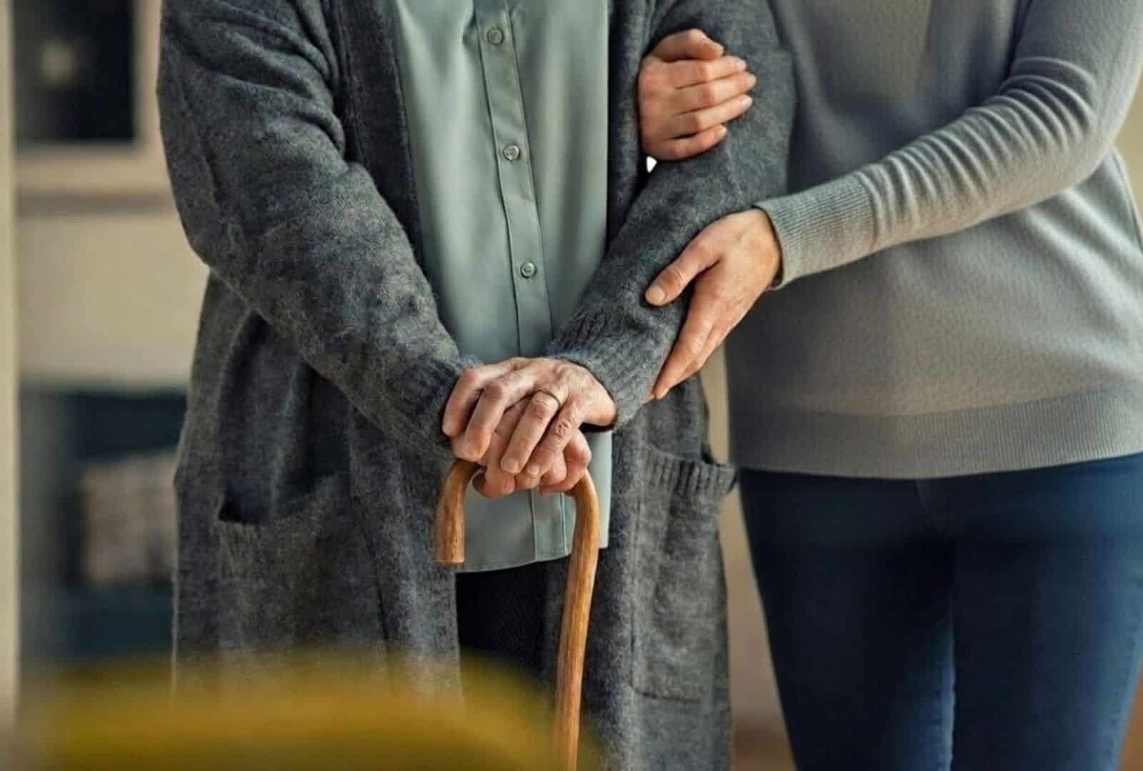 אישה מבוגרת עם קיצור גפה משמעותי נתמכת בעדינות על ידי בתה בעת הליכה, אוחזת במקל הליכה, מדגימה מוגבלות בניידות הזכאית לתג חניה לנכה.