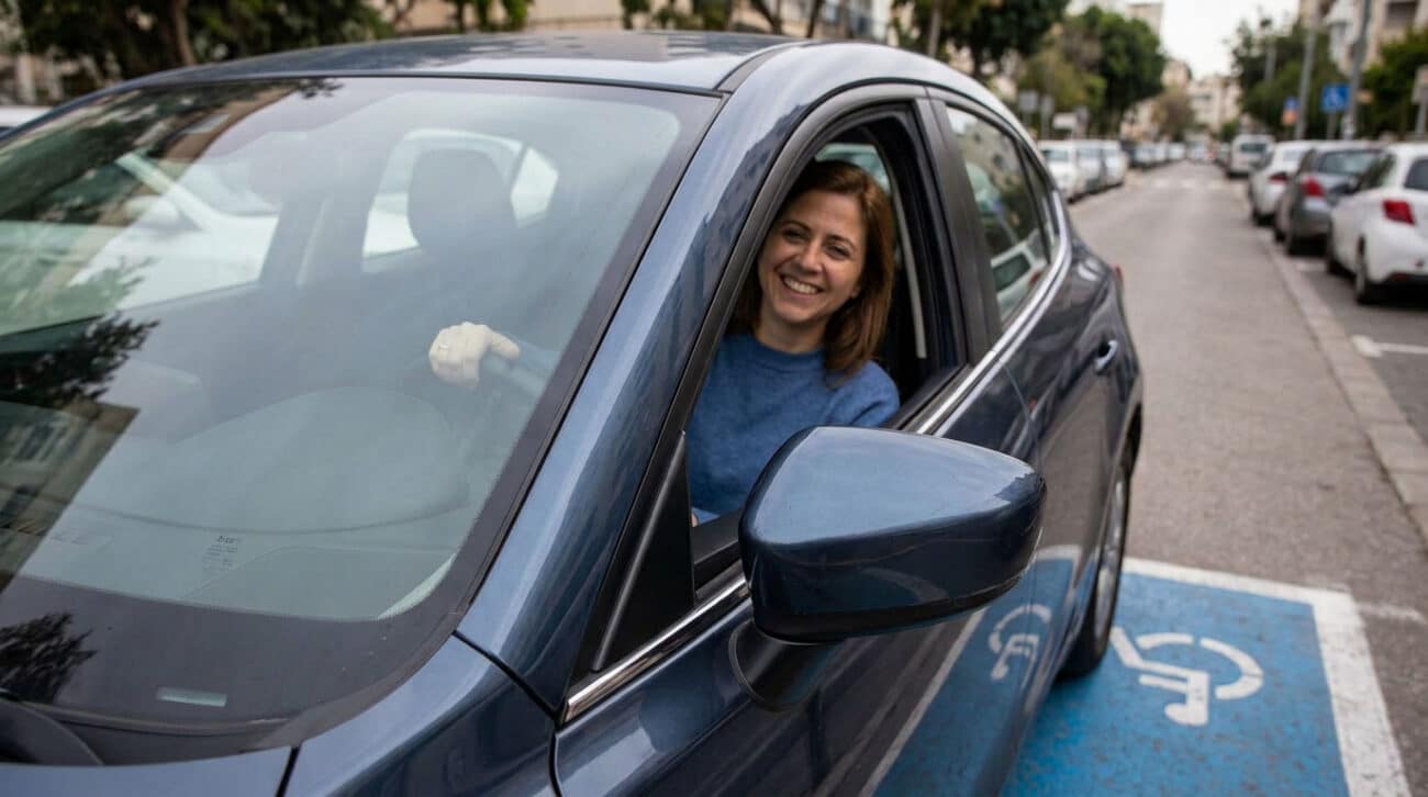 צילום מקרוב של אישה מחייכת היושבת בתוך מכונית כחולה, כשידה על ההגה. מחוץ למכונית, על גבי הכביש, נראה סימון כחול של חניית נכה עם סמל כיסא גלגלים בלבן. המכונית חונה בתוך תחום הסימון ברחוב עירוני.